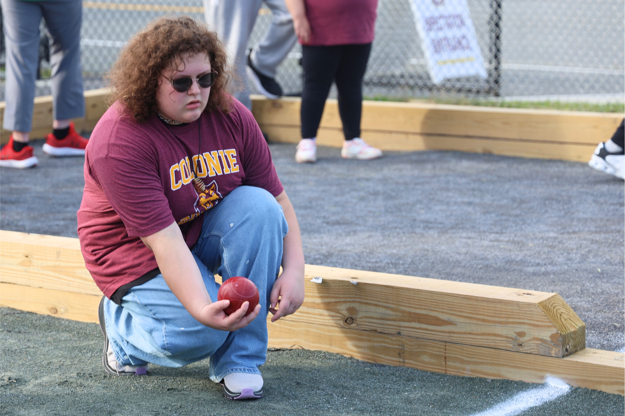 A person with curly brown hair wearing a maroon t-shirt kneels on a gravel court, holding a red bocce ball.
